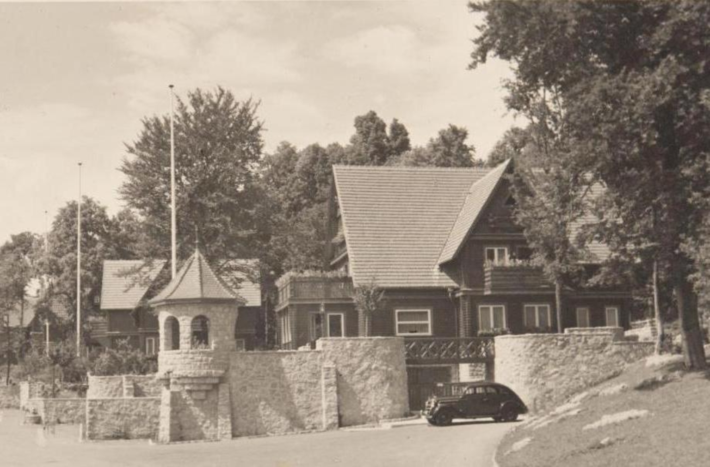 Gesamtansicht der Villa Koch in der Führersiedlung. Das Haus im Heimatstil ist von einer Ziermauer mit Ecktürmchen umgeben. Vor der Einfahrt parkt ein PKW. Im Hintergrund zwei weitere Villen.