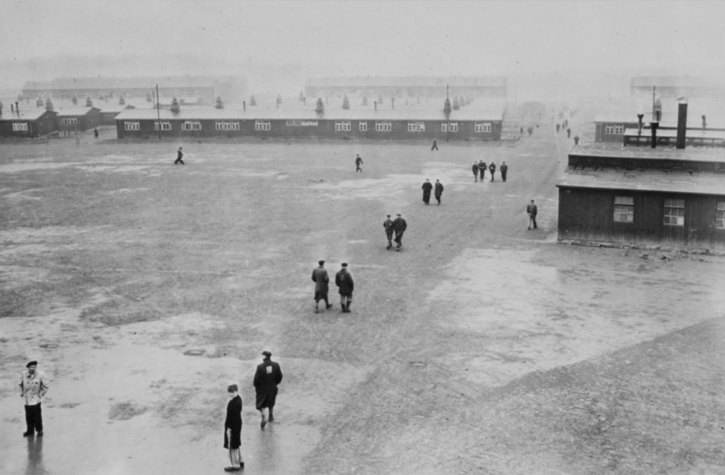 View from the tower of the gate building over the roll call area and the barracks in northern direction. The rear area of the camp disappears in the fog. In the large open area, people can be seen walking around alone or in small groups.