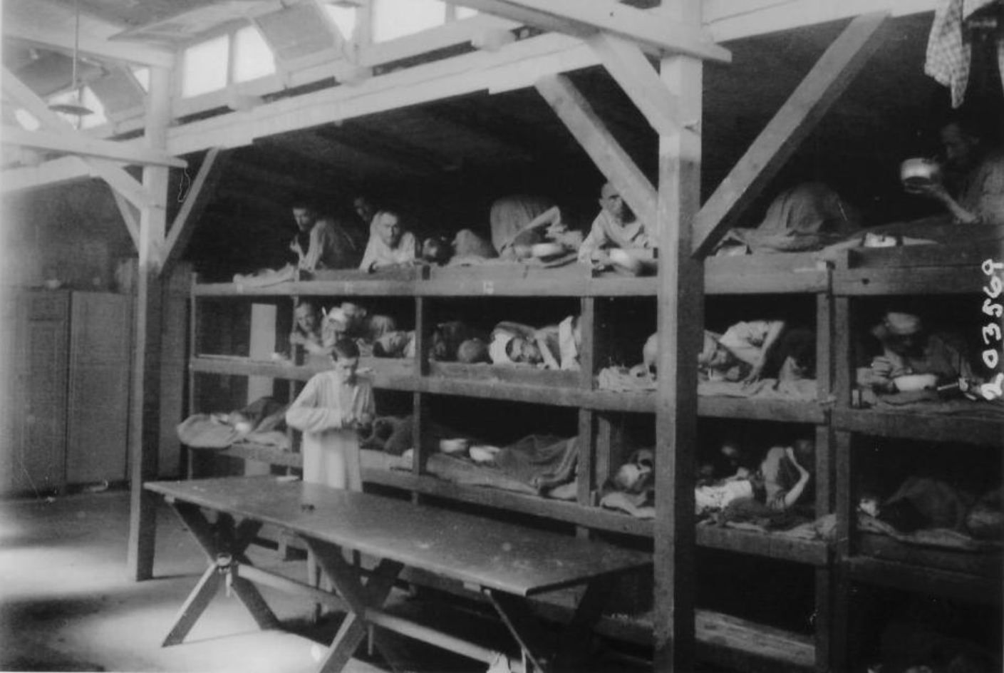 View of Block 61 of the Small Camp. In the background, surviving prisoners in sleeping boxes 11 to 15. These are wooden shelves with four shelves, in which people lie close together with thin blankets. In front of them is an empty wooden table. A liberated prisoner in a long shirt stands next to it