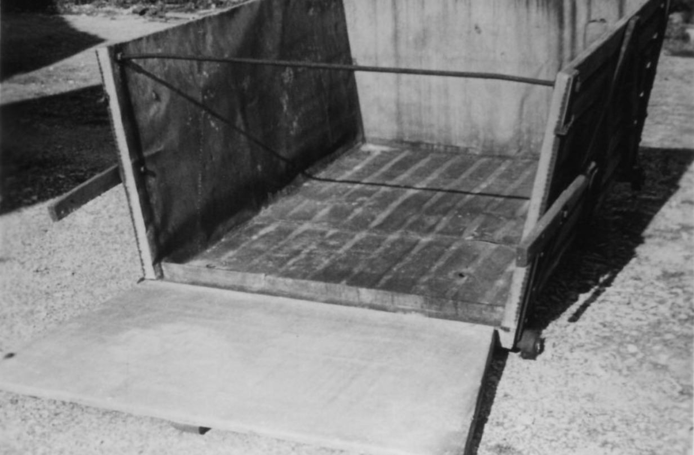 A galvanized trolley for transporting corpses, the front of which is folded out. The trolley is reminiscent of a large mining lorry: it is a large, angular tub, the front of which can be folded open. it stands on small wheels. 