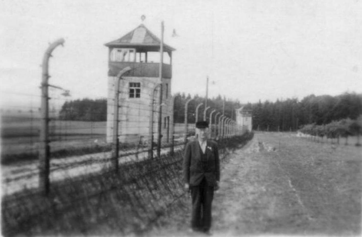 Der befreite Häftling Karl Herrmann steht auf der Innenseite am äußeren Lagerzaun. Hinter ihm der Zaun, dahinter zwei Wachtürme.