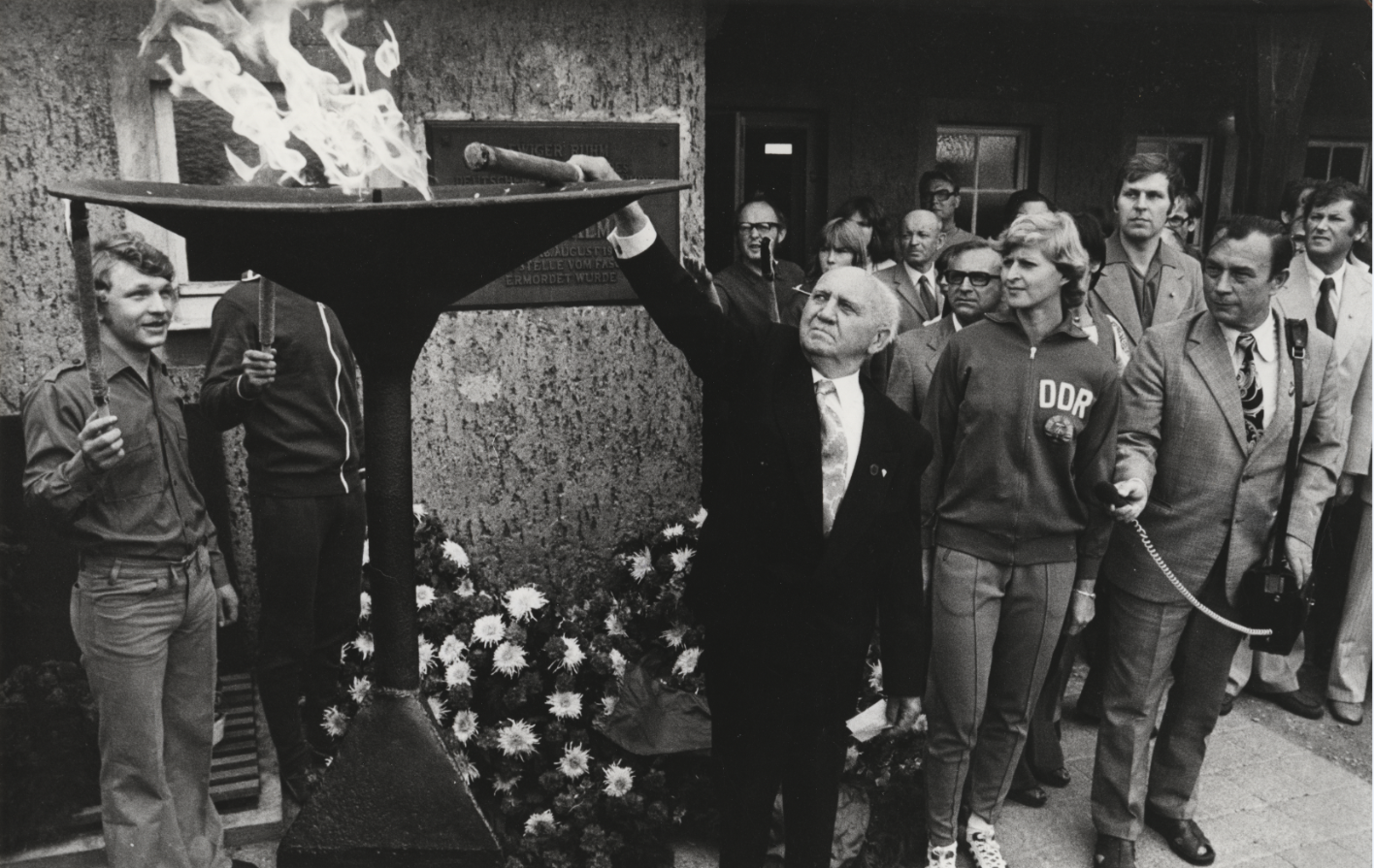 A fire bowl can be seen in front of the Ernst Thälmann memorial plaque, which is being lit by an older man in a black suit. Another man is holding a microphone for him. Behind him are other participants in the event. Two young people stand behind the fire bowl and hold torches. 