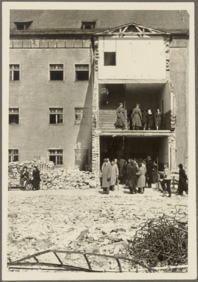 The left wing of the former chamber building can be seen. The staircase is missing the outer wall on the side facing the photographer. The opening is used on the first floor by a larger group of people as an entrance to the building. People are climbing up and down through the stairwell. Through the opening on the second floor, two NVA soldiers can be seen running down the stairs. To the left of the staircase and in the foreground of the picture, remains from the demolition of the camp can be seen.