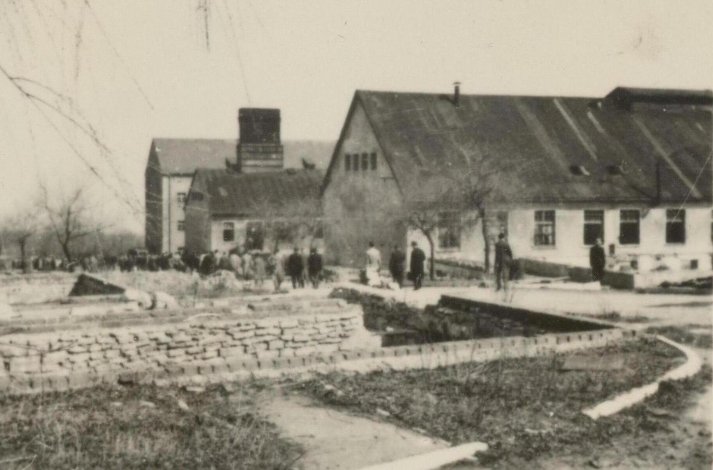 The photo shows, projecting into the picture from the left, three elongated buildings standing one behind the other. A slightly sloping path leads past them, groups of people in civilian clothes are walking on it. The two buildings in front have one story and a high gable roof. The rear building has three stories. 
