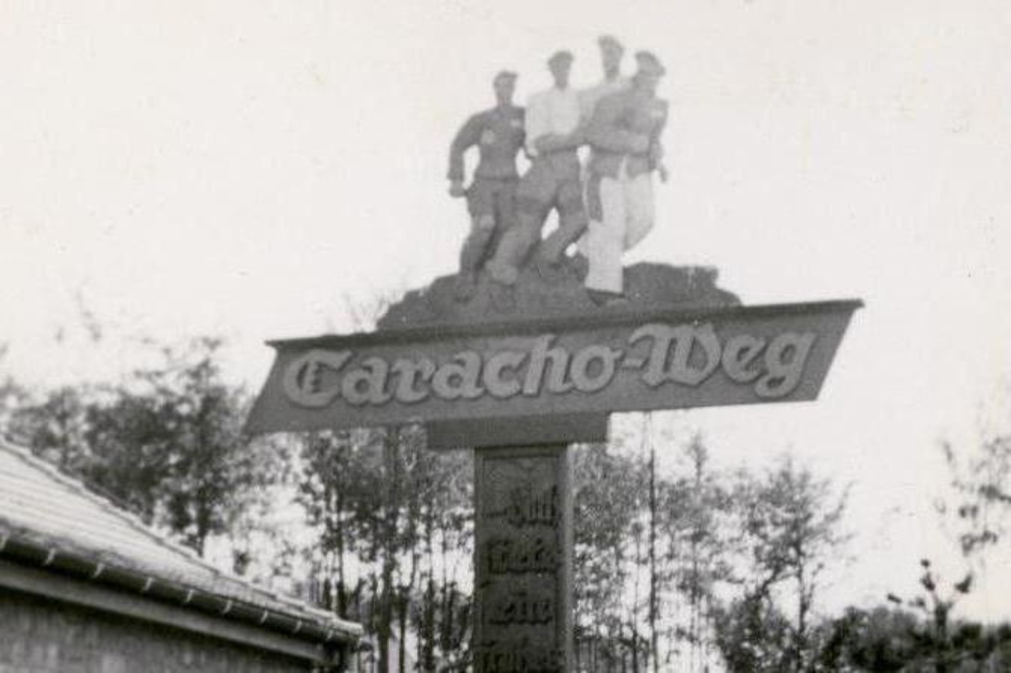 Signpost "Caracho Way". Above the writing is a carved group of figures on the run, driven by an SS man.
