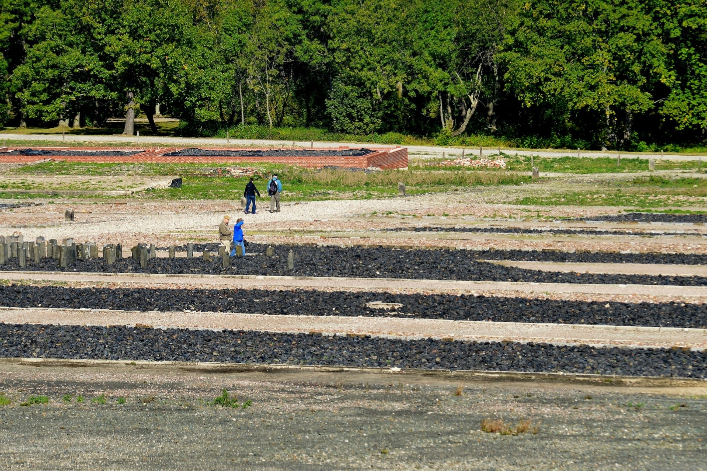 View over part of the memorial site. Dark gravel highlights the outlines of the former barracks. A few visitors.