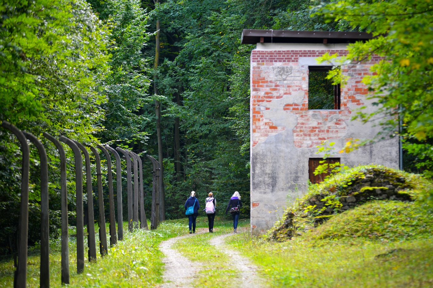 Remains of the camp fence and a watchtower. Visitors walk along a path between them. 