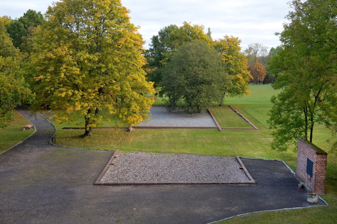 Foundation walls of the former isolation barracks. The gravel surface is interrupted by grass. On the left an intact wall with a memorial plaque