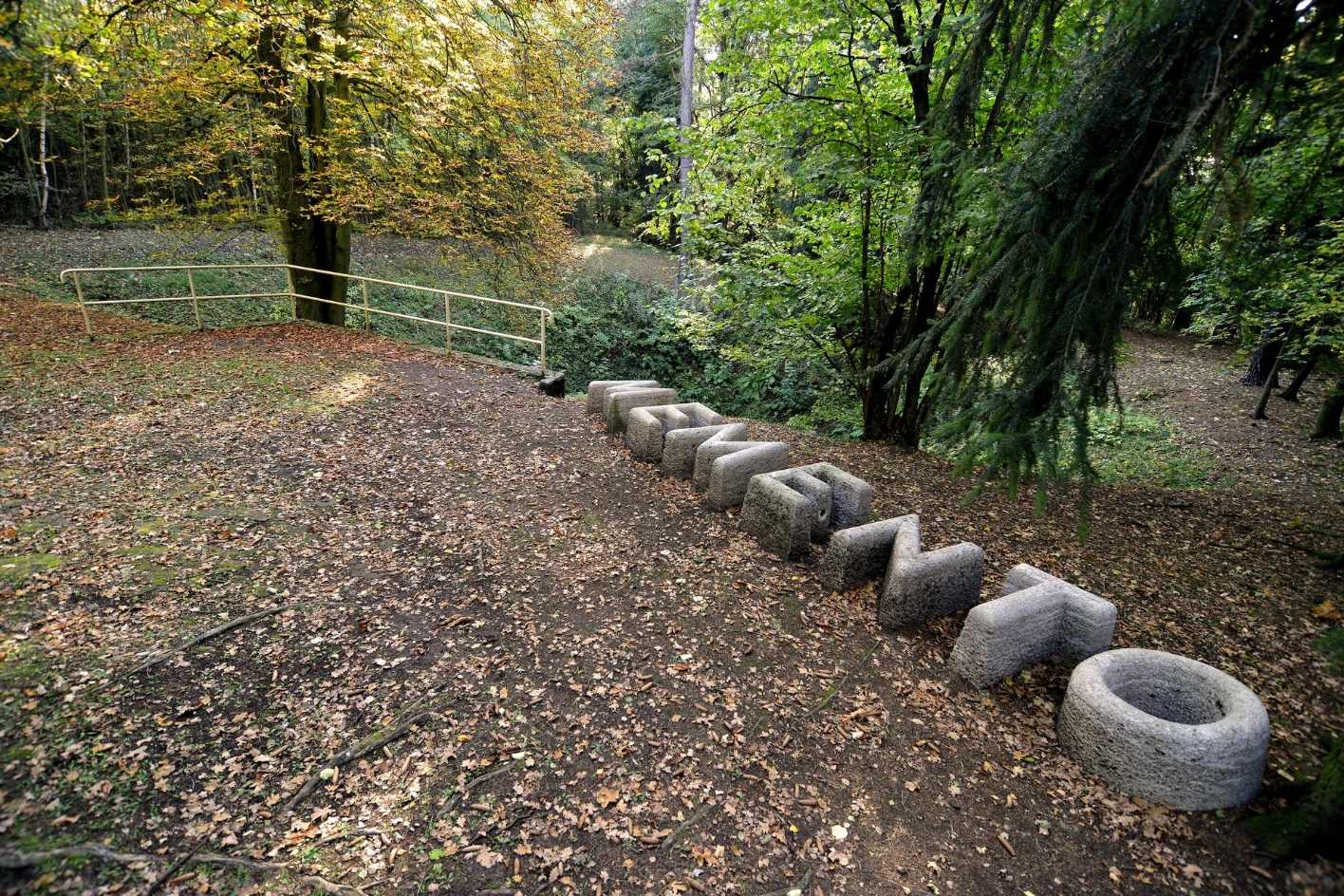 Eine Reihe aus Betonbuchstaben, die das Wort Memento ergeben. Im Hintergrund markiert eine Balustrade einen Erdtrichter, umgeben von Bäumen