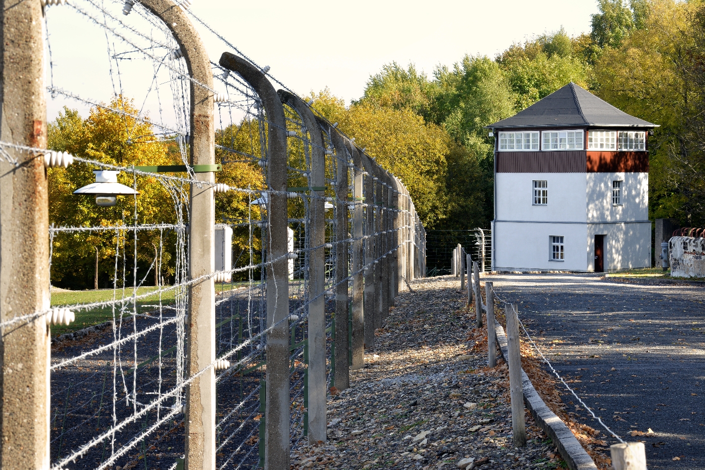 Der Lagerzaun besteht aus Betonpfeilern, Stacheldraht und Elektroleitungen und ist durch einen schmalen Schotterstreifen von einem weiten niedrigeren Stacheldrahtzaun getrennt. Im Hintergrund einer der Wachtürme.