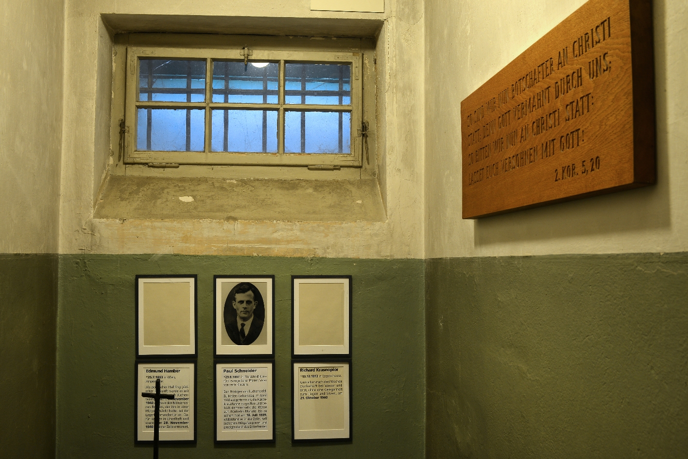 View into a cell of the detention cell building. The cell is very small. Below a barred and blinded window are picture frames commemorating Edmund Haber, Paul Schneider, and Richard Krasnopior. On the left of the wall hangs a wooden plaque with the engraving: "So now we are ambassadors in Christ's stead, in that God admonishes, as it were, through us; we pray for Christ: be reconciled to God!" 