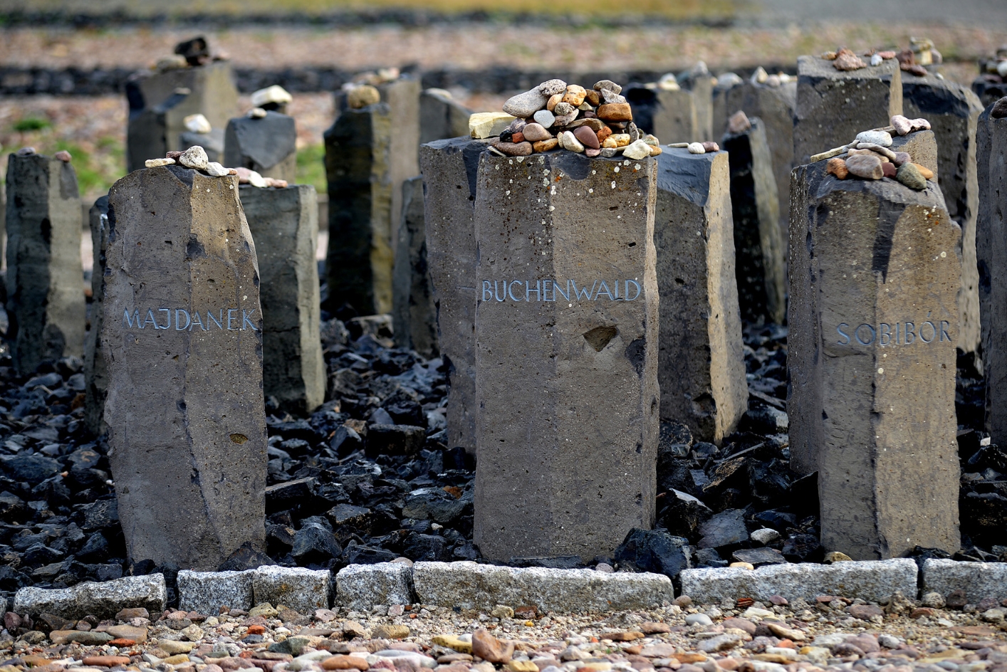 Columnar, waist-high stones set in a black siltstone field. On the top of the stones were deposited sporadically small stones. The three stone columns in focus are engraved:. Left: "Majdanek", center: "Buchenwald". Right: "Soribor".