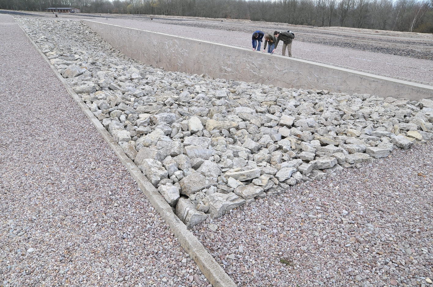 A long rectangular area filled with quarry stones. There are fewer stones towards the center, which is why a depression can be seen. In the background are three visitors, each of whom places a small stone on the edge of the depression. 