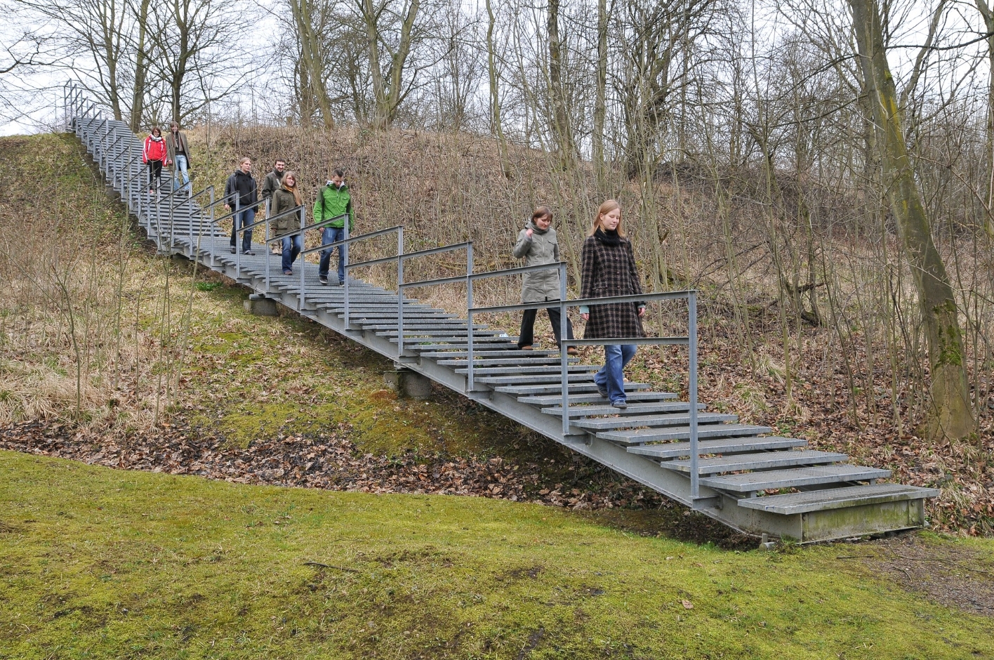 Besucher:innen begehen die Zeitschneise vom Lager aus. Eine Treppe führt zum ehemaligen Postenweg hinab.