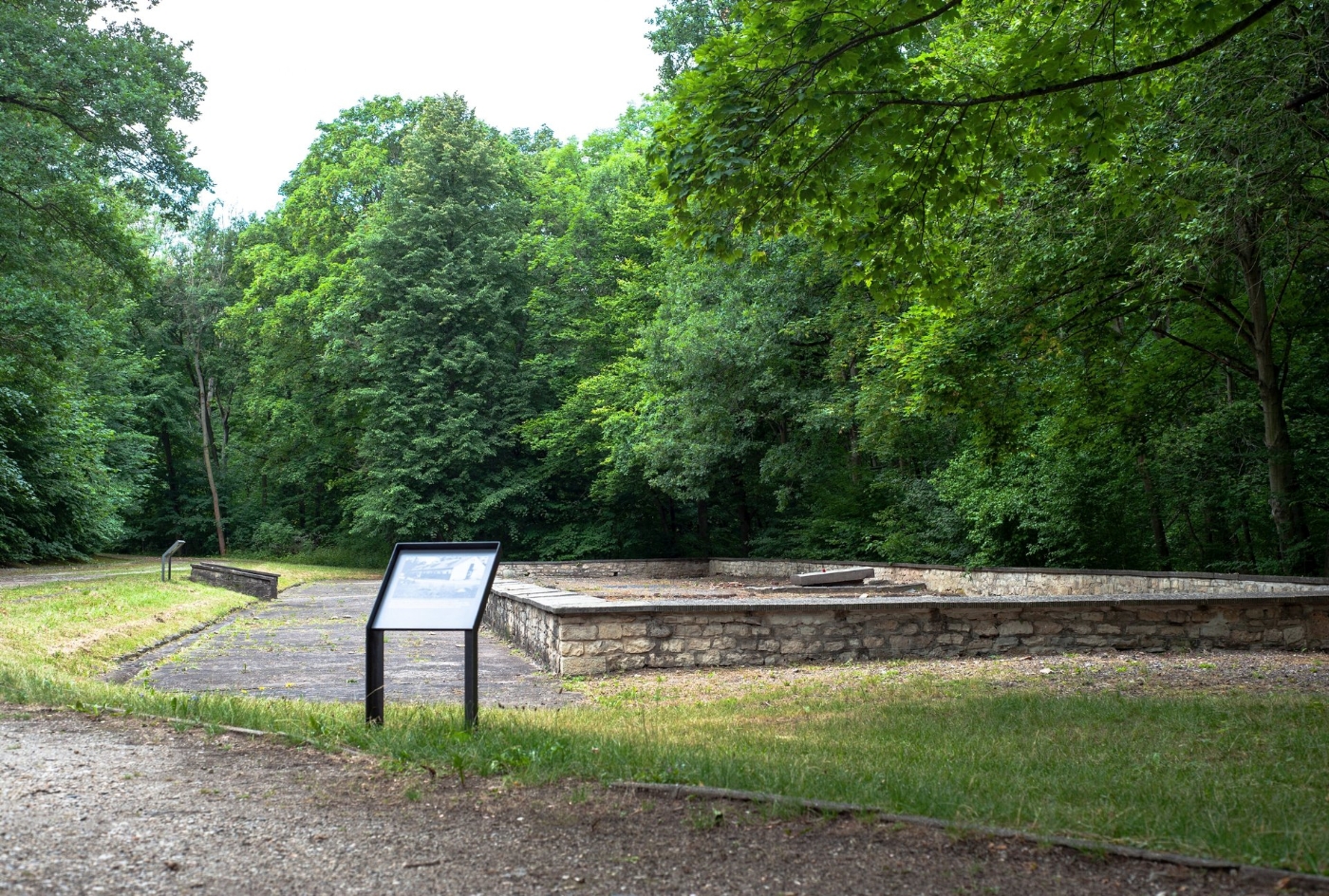 In the foreground, an info sign indicating that this is the site of the former horse stable. Further back the highlighted foundation walls of the stable, in the center of which is a memorial stone.