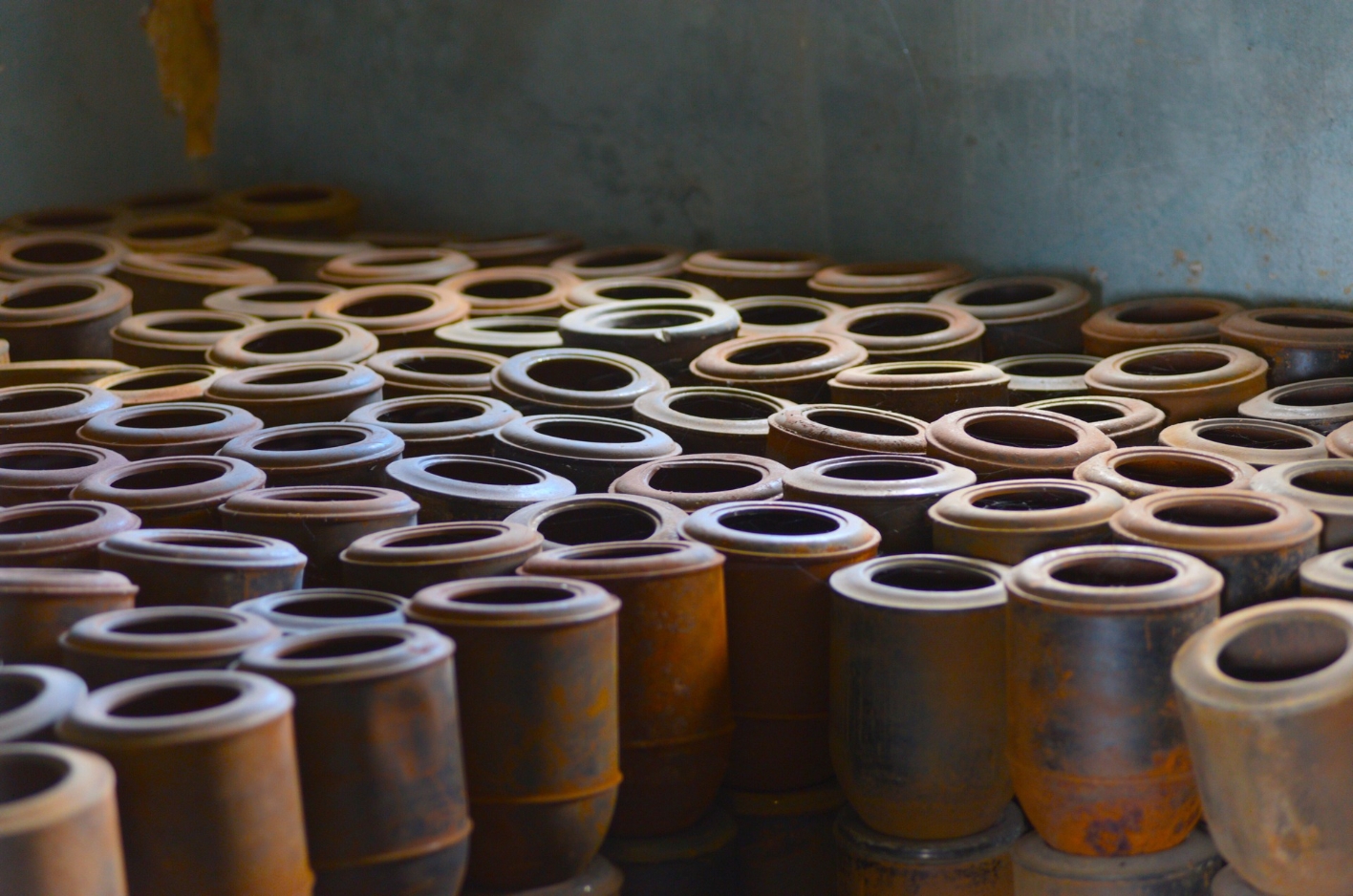 A room, the floor of which seems to be covered with plain, metal urns, which are placed close to each other. Rust is visible on many of the containers.