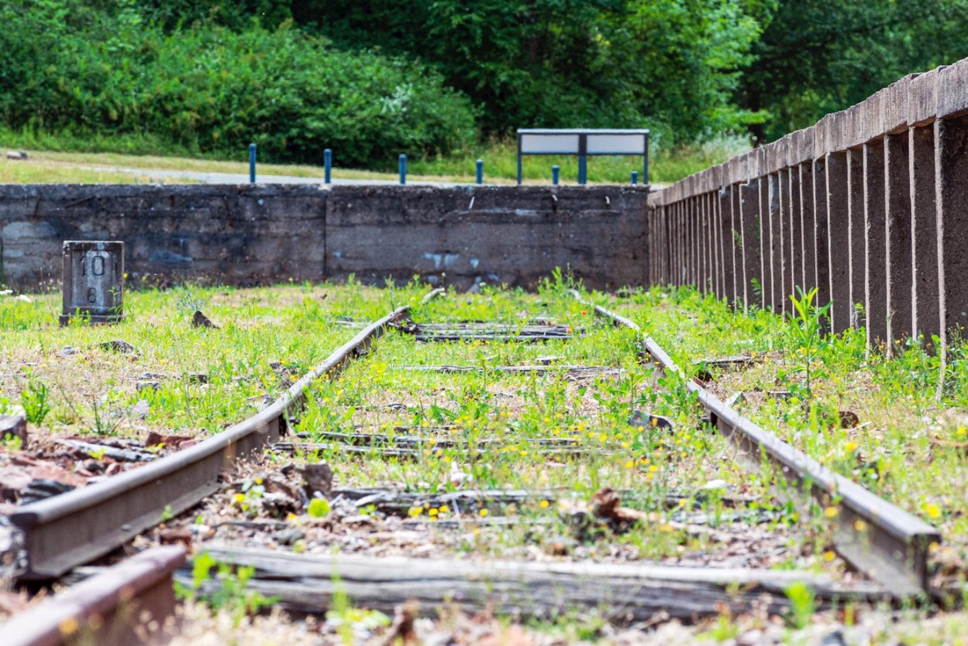 Froschperspektive vom Gleisbett aus. Die Schienen rechts vom hohen Bahnsteig sind verbogen. Zwischen ihnen ragen Graeser hervor. Links ist ein Meilenstein der Buchenwaldbahn zu erkennen.