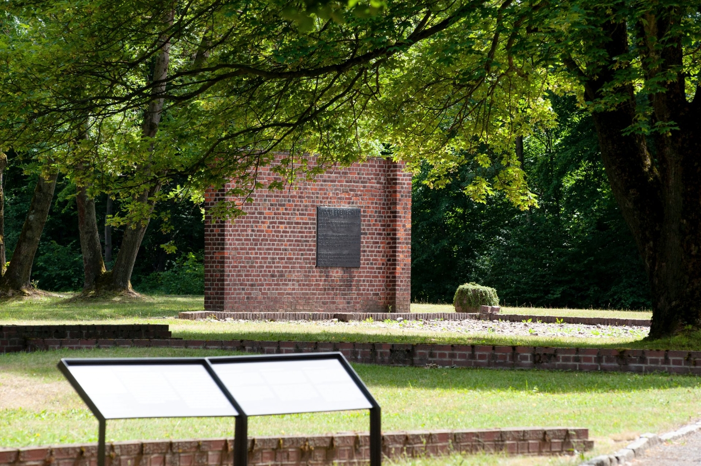 View of the memorial wall with plaque of the Rudolf Breitscheid Memorial. In front of it, the remains of the foundations of the former isolation barracks can be seen. In the foreground an information sign.