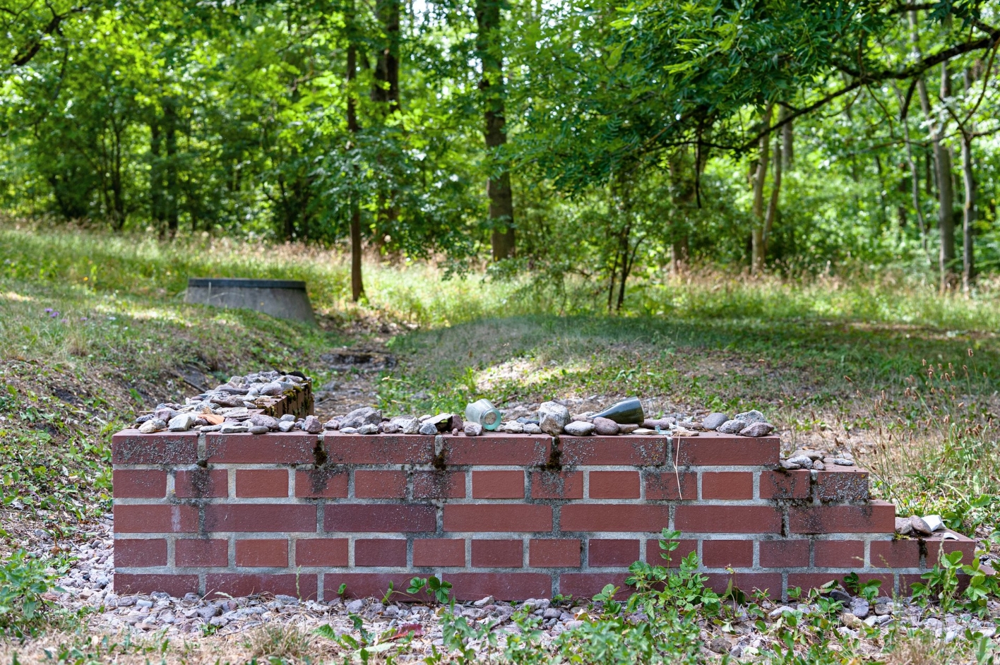 A small, L-shaped brick wall remnant. On the low remains of the wall are deposited individual finds such as stones or parts of bottles.