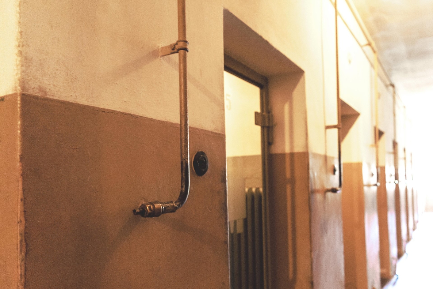 Eine Reihe von Zellentüren. Neben jeder Tür ein dünnes Metallrohr, das zur Heinzungssteuerung gehört. Darüber an der Wand ist das Drehrad der Heizungssteuerung angebracht.