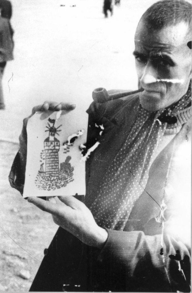 A liberated prisoner holds a piece of tattooed human skin with a lighthouse on it up to the camera.