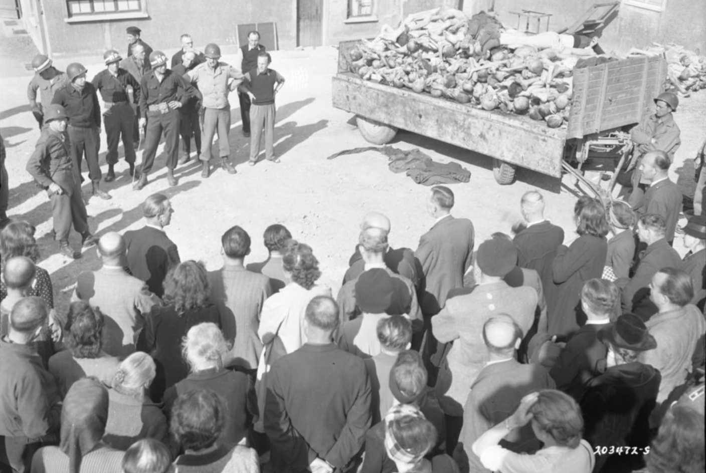 Residents of Weimar during the tour of Buchenwald concentration camp ordered by the Americans in the courtyard of the crematorium in front of a truck trailer loaded with corpses. To the left of the trailer, wearing a steel helmet and light-colored jacket, the American commander Lorenz Schmuhl.
