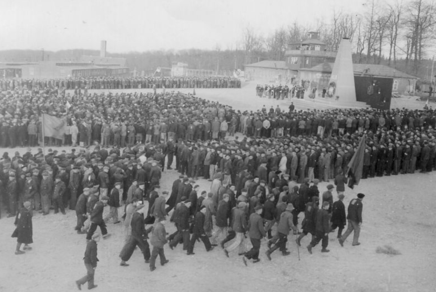 Befreite Häftlinge bei der ersten Gedenkfeier für die Toten des KZ Buchenwald. Der Appellplatz ist dicht an dicht mit Menschen gefüllt. Im Hintergrund ist das provisorische Mahnmal für die Toten. Vereinzelt Fahnen.