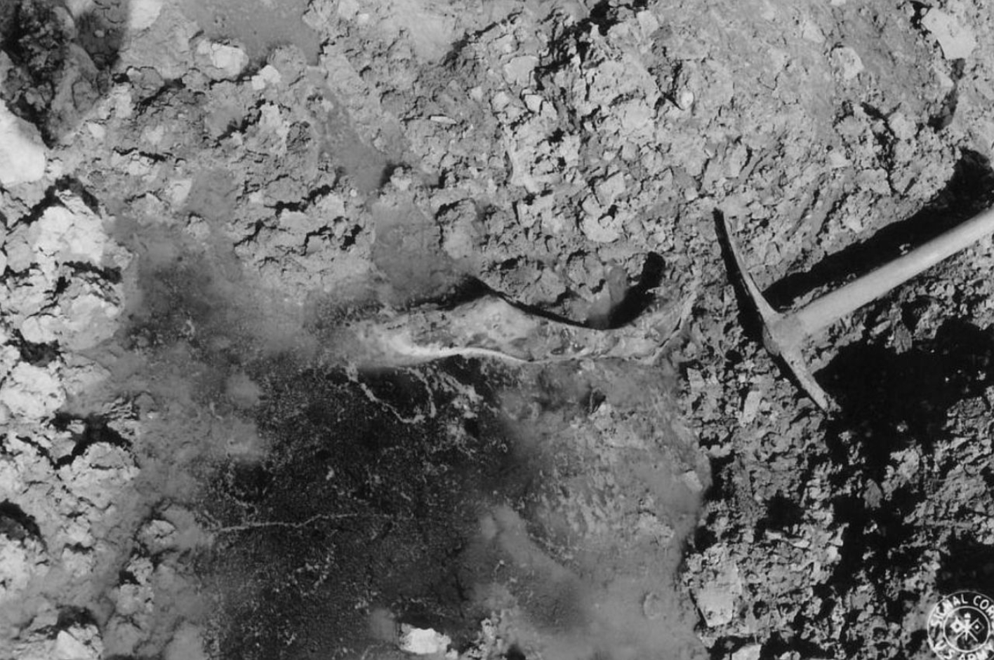 Blick in eines der Massengräber des Buchenwalder Außenlagers Ohrdruf. Aus einem schlammigen Loch ragt ein Bein heraus.