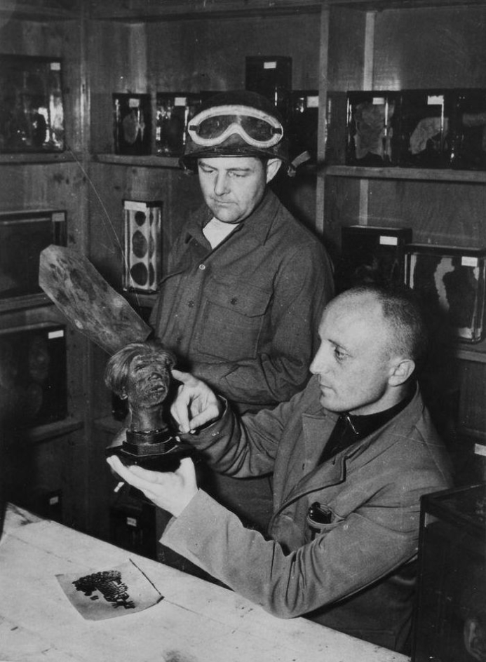 US soldiers can be seen in the pathology department. The one in front is sitting, the one behind is standing. The seated soldier inspects a shrunken head that he has picked up. The soldier behind him is also looking at the shrunken head, but is holding tattooed human skin in his hands.