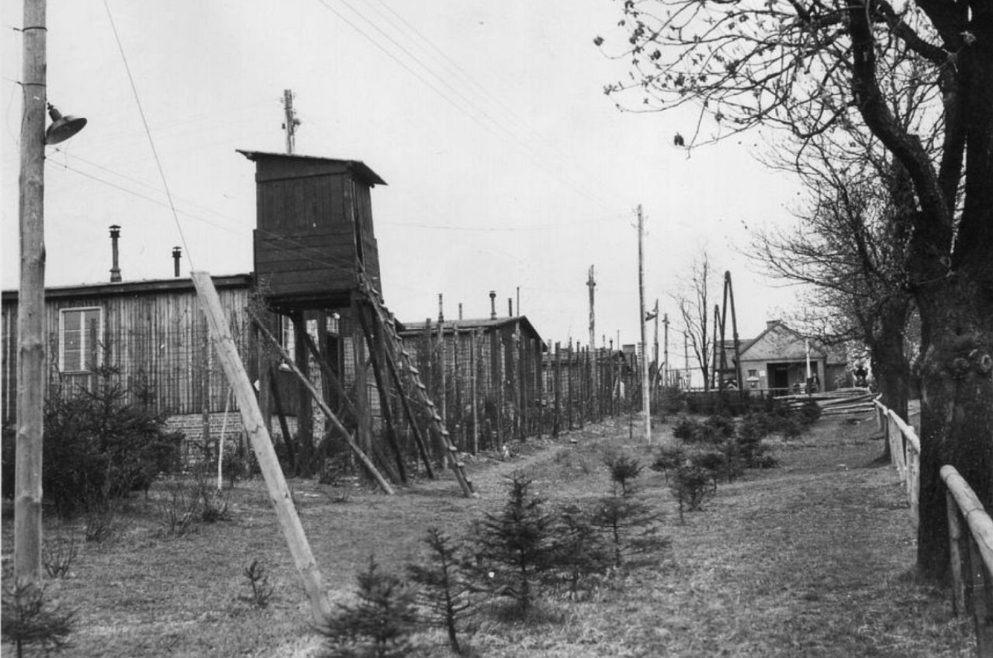Blick auf einen Wachturm und den Lagerzaun des Buchenwalder Außenlagers Ohrdruf. Der Wachturm wirkt provisorisch und erinnert an einen Jägerstand.