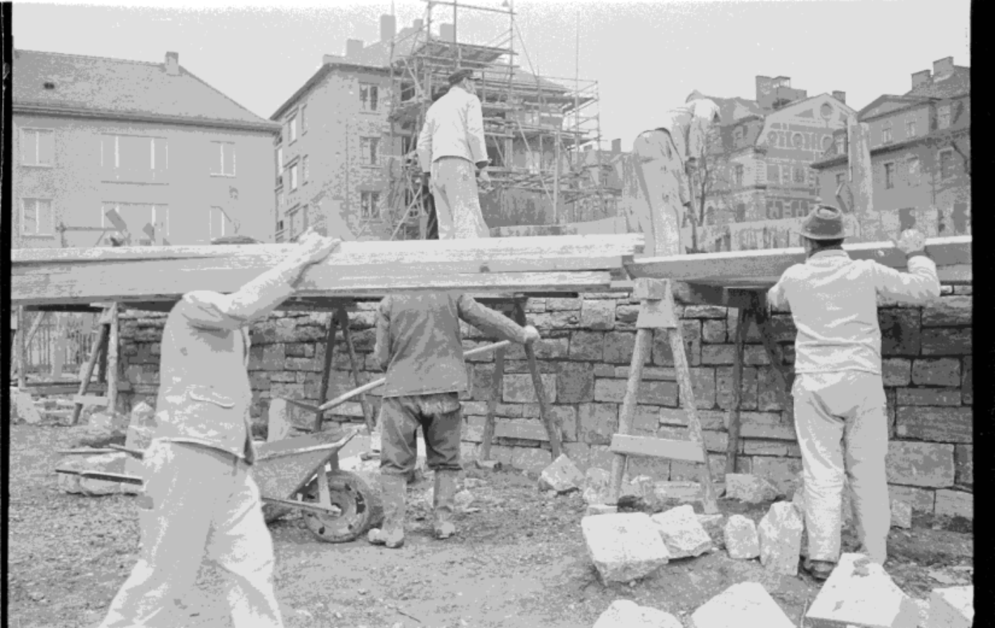 Zusehen sind Bauarbeiter. Links trägt ein Mann Holzlatten von Links nach Rechts. Rechts ist ein Maurer zu Gange. Auf einer Mauer Steht ein weiterer Arbeiter in der Mitte des Bildes. Im Hintergrund sieht man eingerüstete Gebäude.