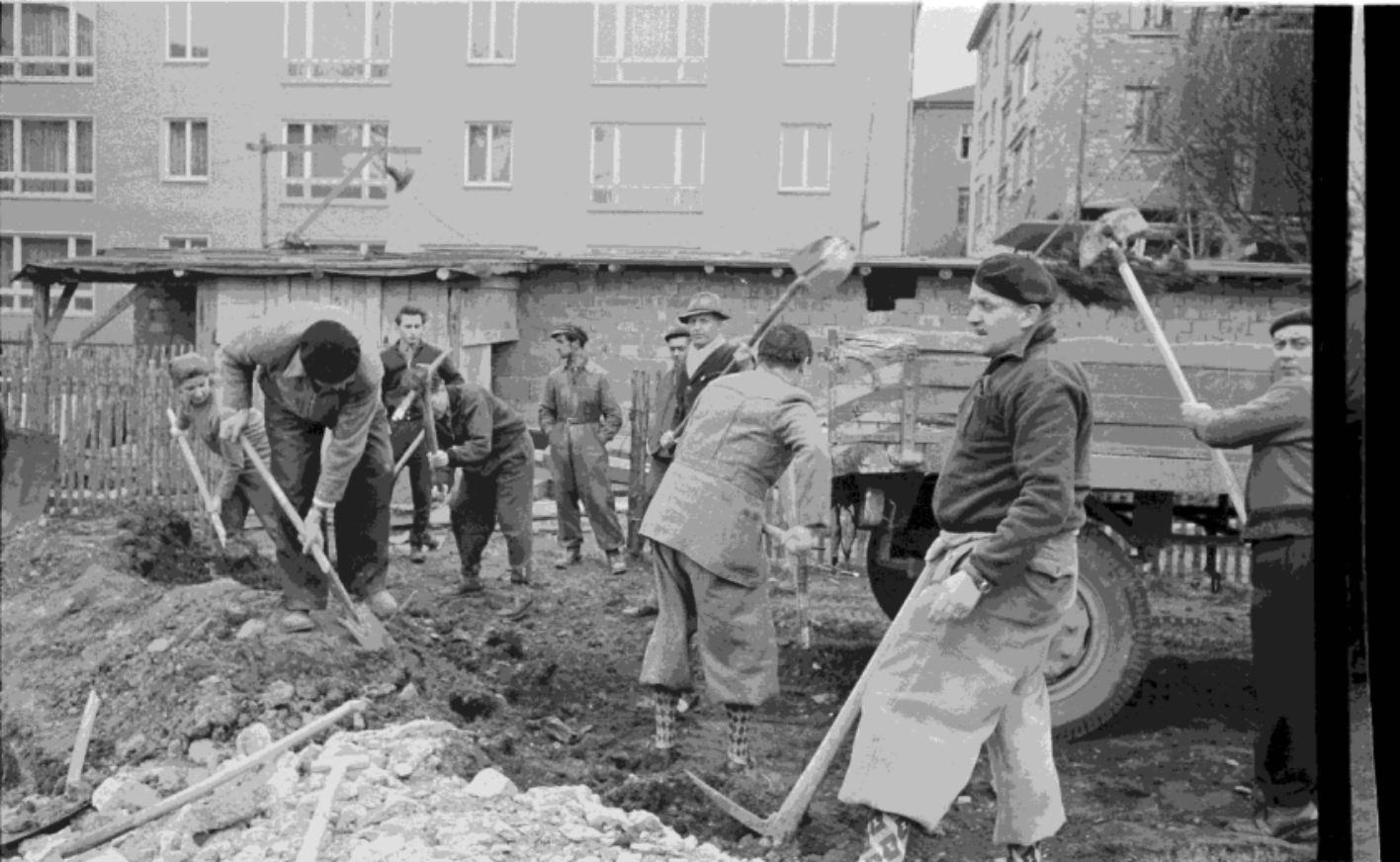 Auf dem Bild ist eine größere gruppe Bauarbeiter zu sehen, die mit Spaten einen Graben ausheben, und den abgetragenen Boden auf eine Fahrzeugladefläche schaufeln.