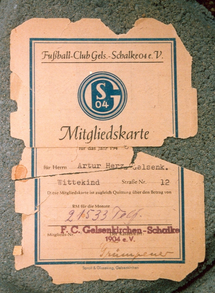 Zusehen ist eine Mitgliedskarte des FC Schalke 04 für Arthur Herz. Sie hat eine lachsfarbende Farbe und ist an den Rämdern eingerißen. Das Schalke 04 Logo ist gut sichtbar in der Mitte der Mitgliedskarte zu sehen.
