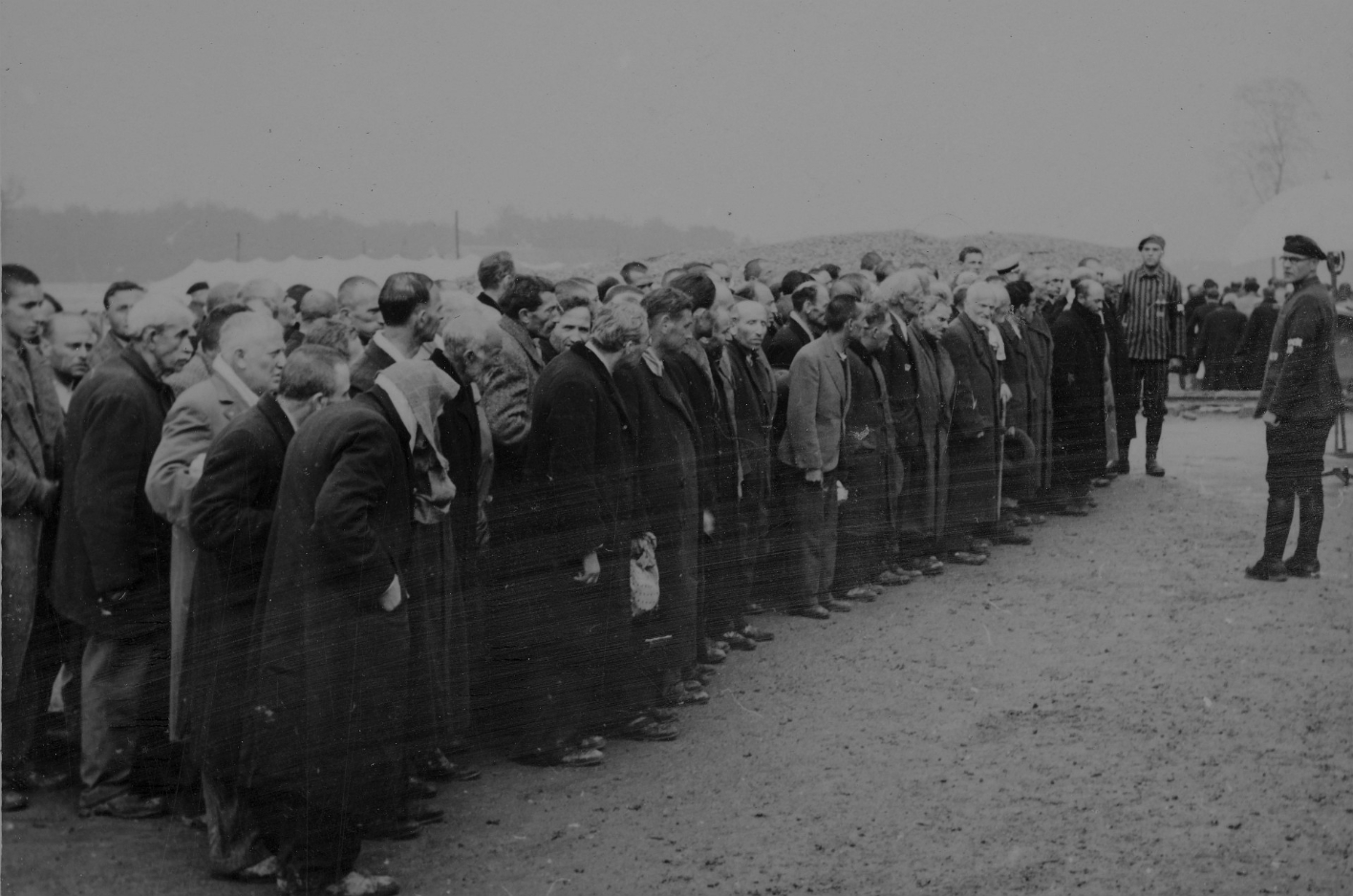 In Zivil angezogene Männer stehen aufgereiht. Sie sehen sichtlich beunruhigt aus. Vor den aufgereiten Männern steht ein Mann in SS-Uniform, der zu ihnen spricht.