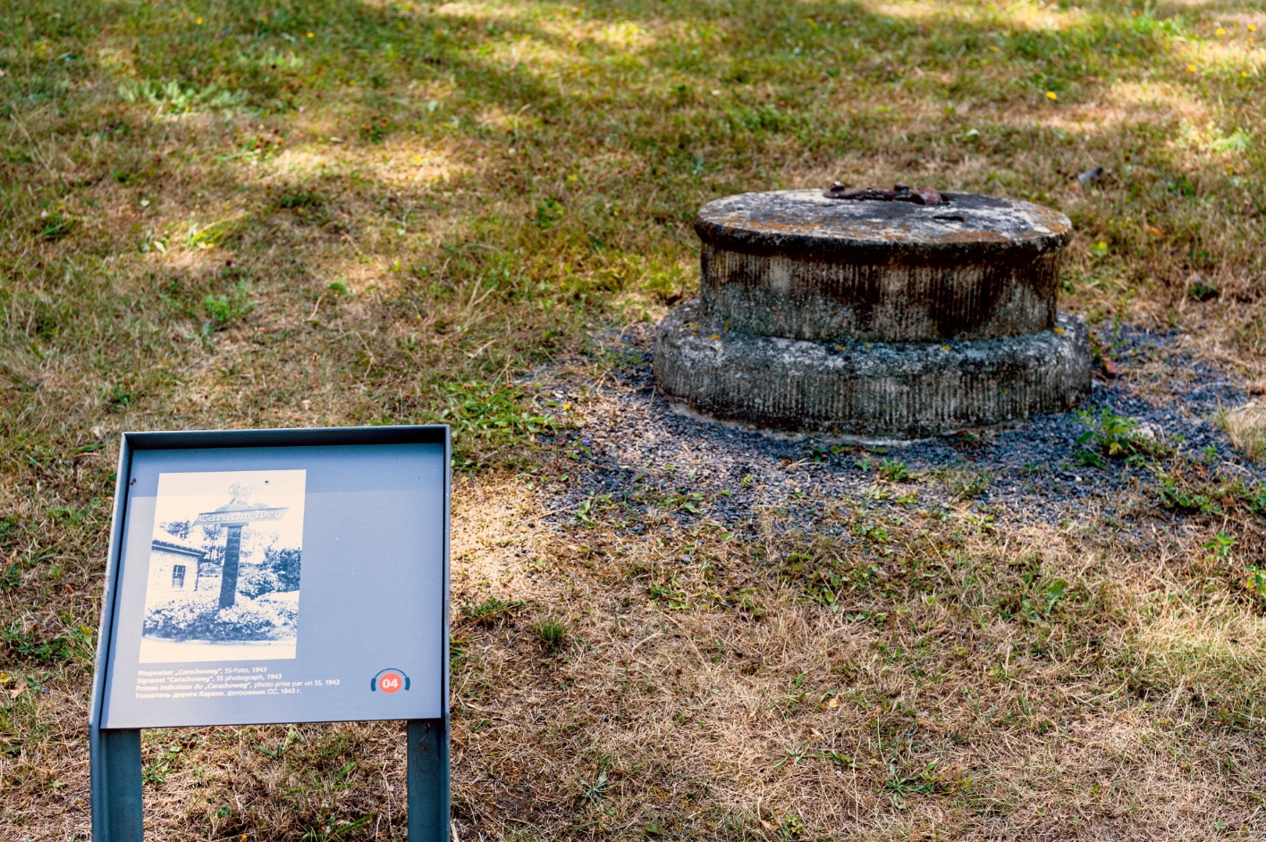 A round concrete base stands on a lawn. The rusty metal envelopes on its top suggest that a wooden signpost was once fixed here.