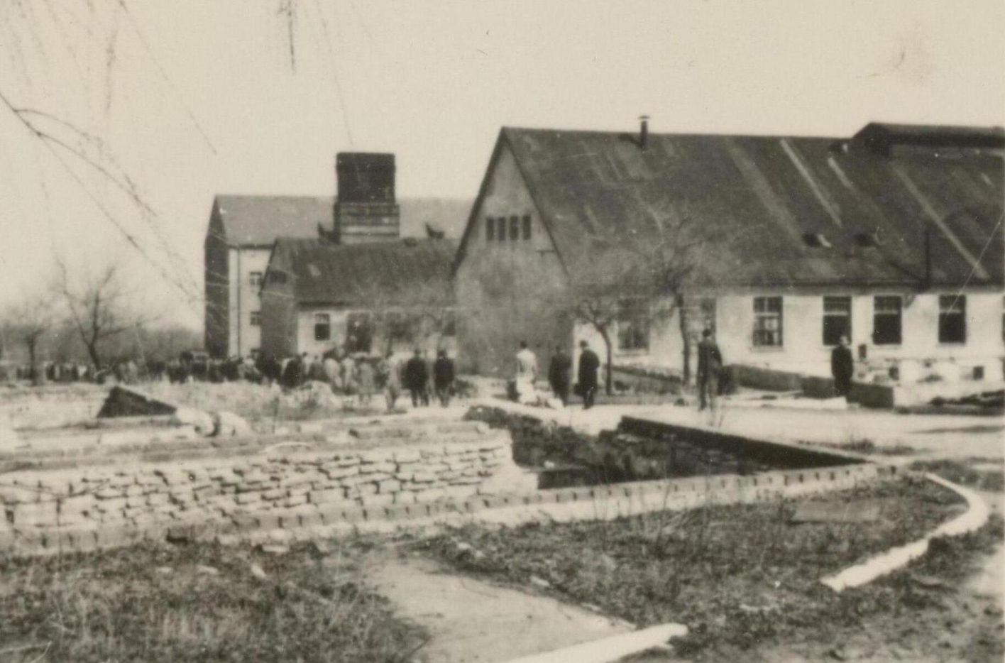 Das Foto zeigt, von links ins Bild ragend, drei längliche, hintereinanderstehende Gebäude. Ein leicht abschüssiger Weg führt an ihnen vorbei, Gruppen von Menschen in ziviler Kleidung sind darauf unterwegs. Die beiden vorderen Gebäude haben ein Stockwerk und ein hohes Giebeldach. Das hintere Gebäude hat drei Stockwerke. 