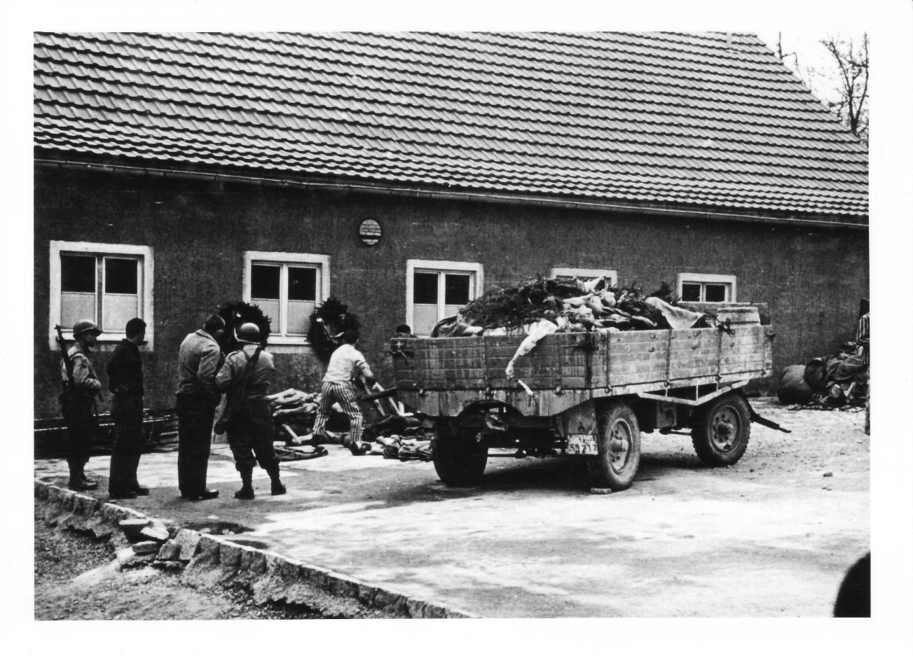 U.S. Army personnel and liberated prisoners in front of a truck trailer loaded with bodies of deceased prisoners in the crematory courtyard.