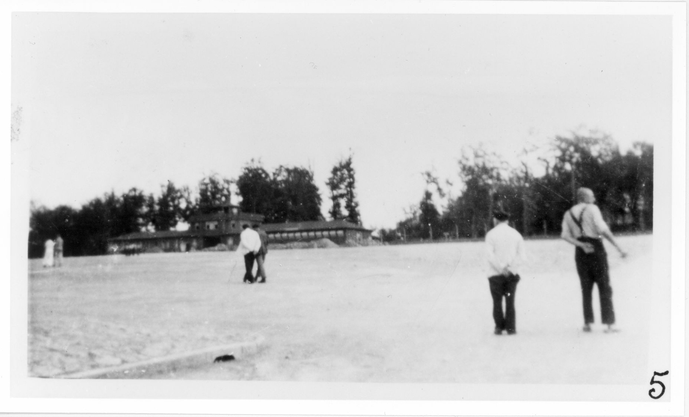 Prisoners walk in small groups or in pairs at a great distance from each other on the roll call square. The gate building in the central background.