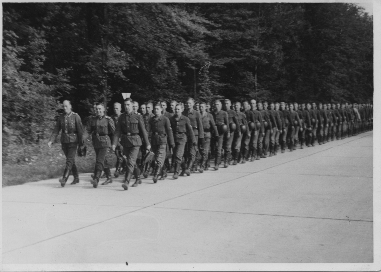 SS-members of a recruit unit of the SS-Totenkopfstandarte 14 marching back after their swearing in on the Blutstraße. In rows of three, a long line of uniformed men walks along the concrete slab road through the forest. 