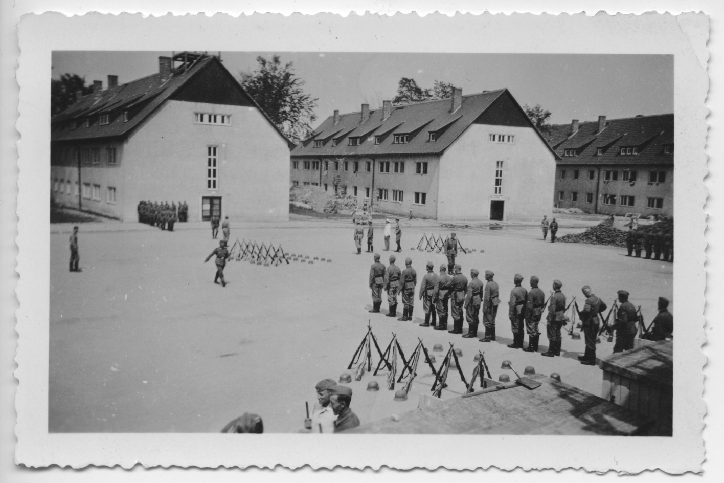 SS members of Totenkopf-Standarte 14 during drill exercises on the parade ground. The men are standing in rows. Rifles are leaning against each other on the ground and helmets are lying next to them. Three Hundertschaftskasernen can be seen in the background.