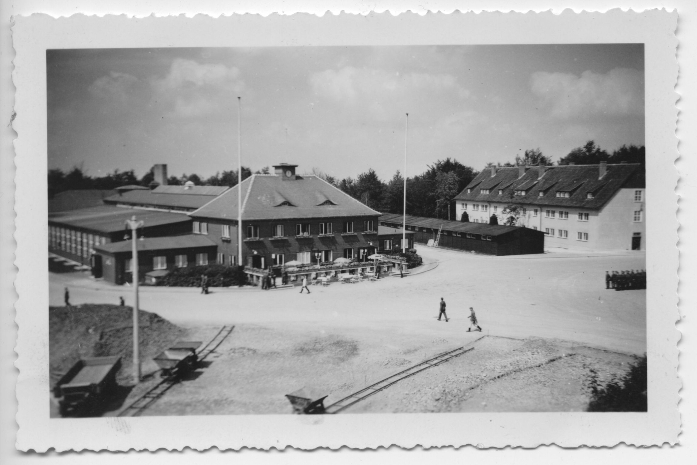 Blick über den Exerzierplatz auf das SS-Kasino. Vereinzelt sind SS-Männer zu sehenn die über den Platz laufen. Im Vordergrund sind 2 Loren auf Scheinen erkennbar.