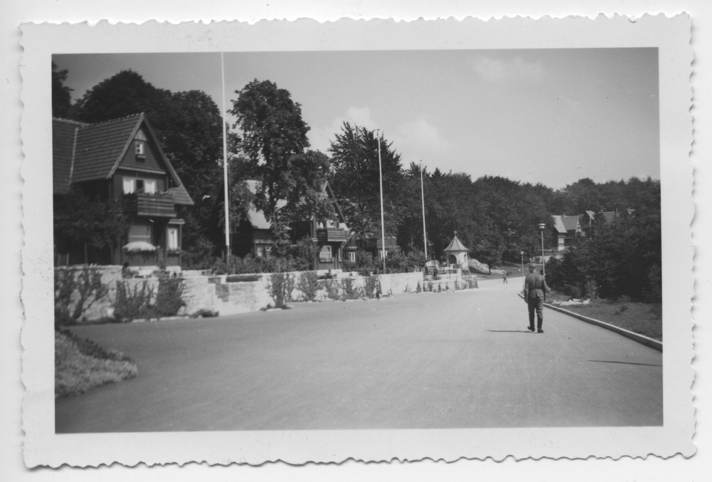 Blick auf die Villenstraße in der SS-Führersiedlung, 1940. Die Straße ist breit, die zweistöckigen Villen mit Balkonen zur Straße stehen durch hohe Bäume voneinander getrennt.