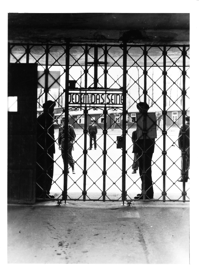 Blick durch das Lagertor auf den Carachoweg und die dahinterliegende ehemalige SS-Adjutantur. Hinter dem Tor stehen befreite Häftlinge und amerikanische Soldaten. Der Schriftzug am Lagertor "Jedem das seine" ist von der Seite des Fotografen lesbar.