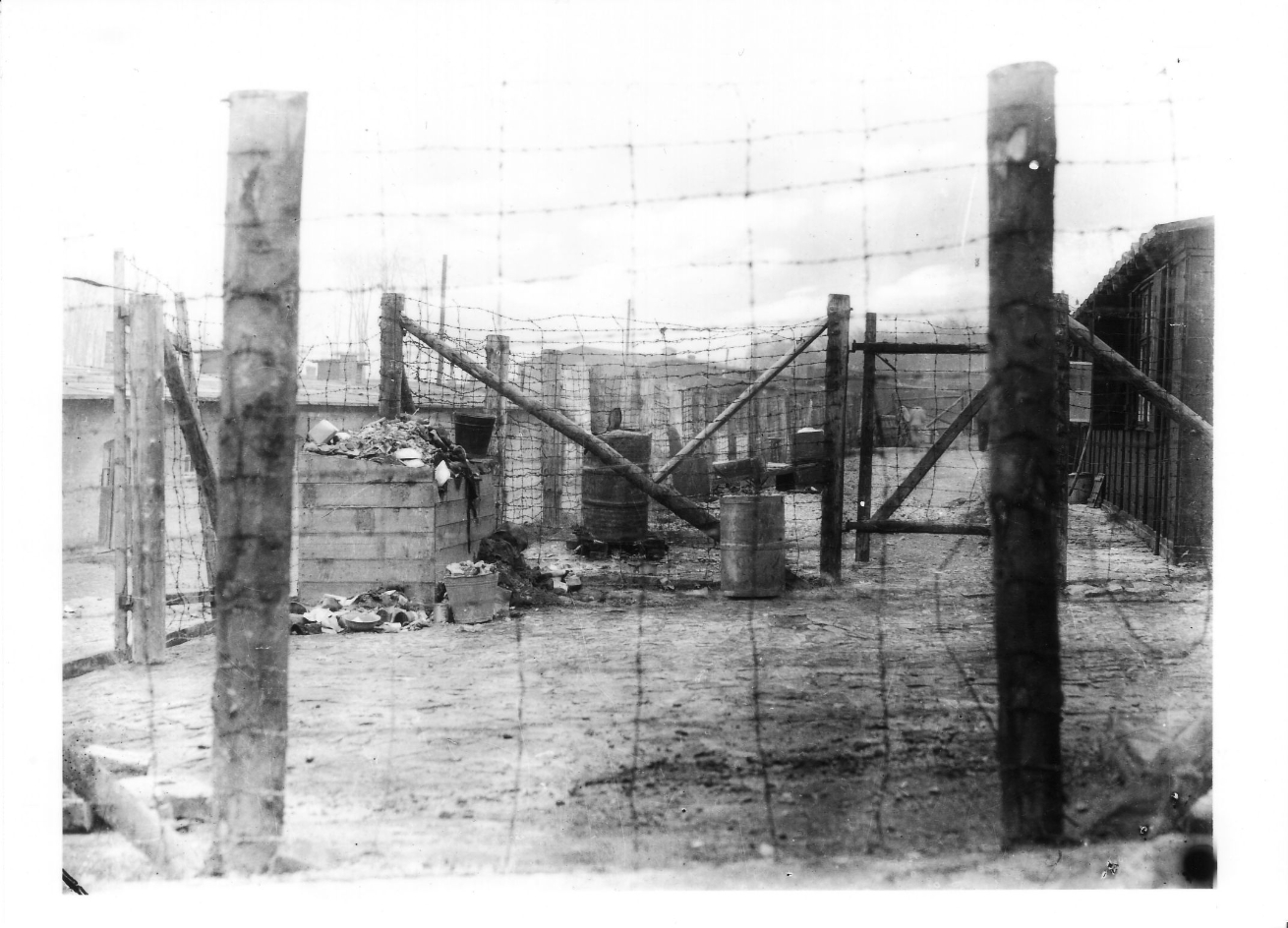 Block 54 on the right, Block 60 in the background, the latrine building on the left. A container full of garbage and several barrels. Photographed through a simple wire fence made of rough beams.
