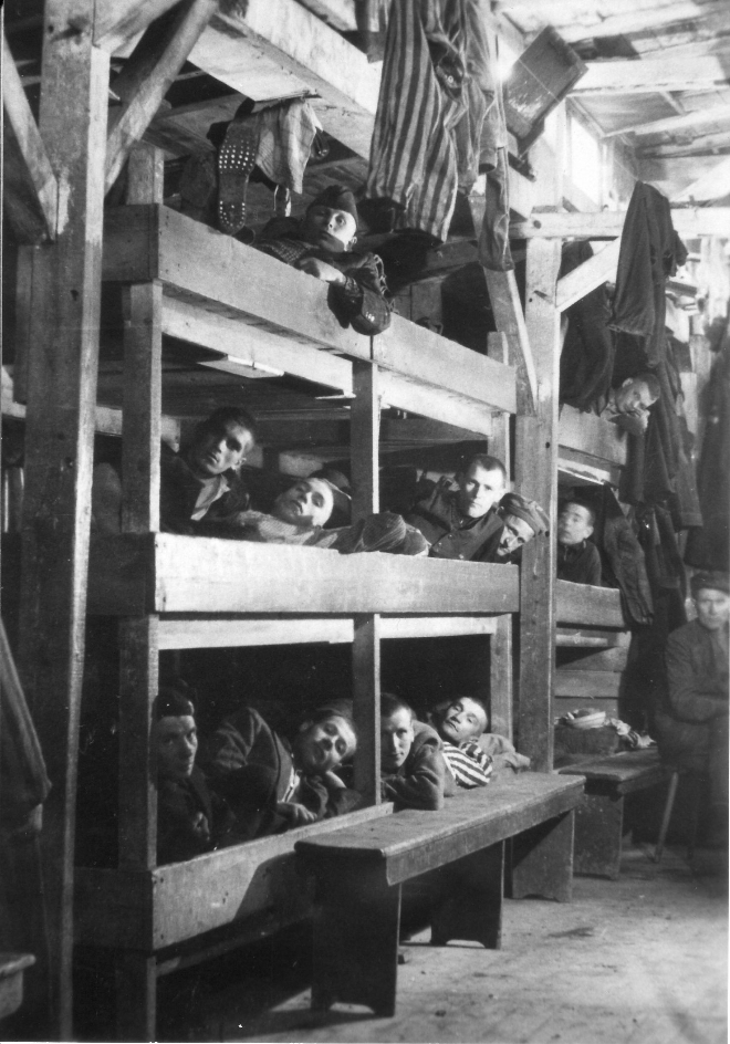 Several four-story cots can be seen, which are occupied several times. The liberated prisoners lying in them look out of their cots into the camera.
