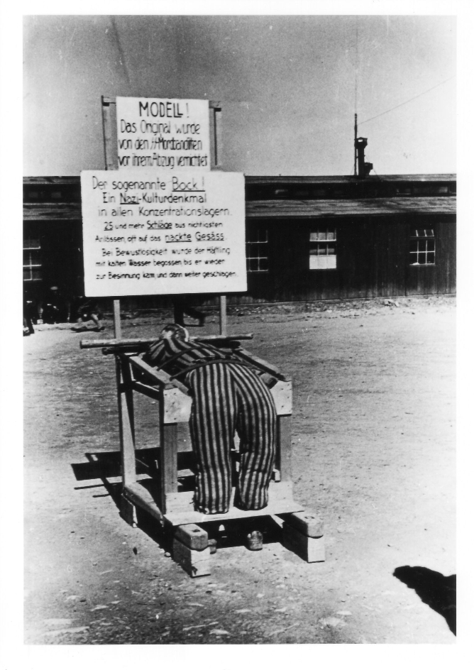  Rekonstruktion des Prügelbocks mit einer Häftlingspuppe auf dem Appellplatz. Darüber sind zwei Schilder angebracht, die Bestrafungspraxis im Lager erklären.