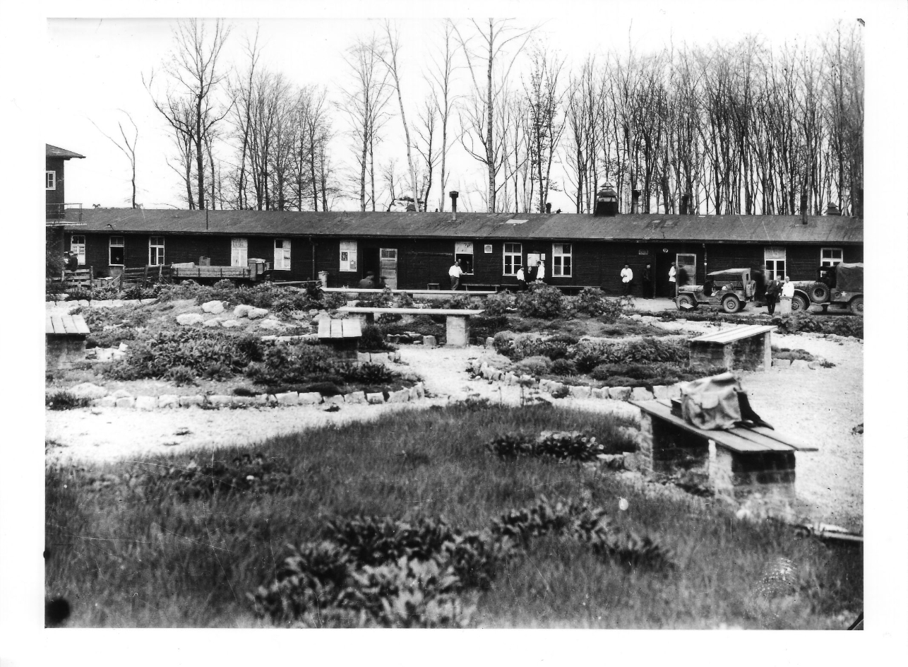 The garden area of the prisoners' hospital, which is characterized by idyllic flower beds, benches and branched paths. In the background is the long low-rise building of the prisoners' hospital. In front of it are isolated nursing staff with white jackets.