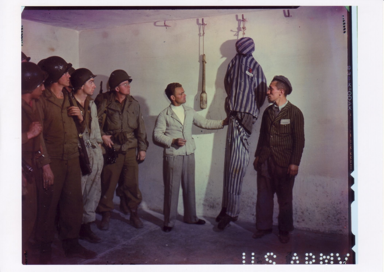 On the left is a group of American soldiers watching the demonstration. In the center of the picture, a former prisoner stands in front of a doll in prisoner's clothing hung on a wall hook and makes an explanatory gesture. On the right, another former prisoner is speaking to the American soldiers.