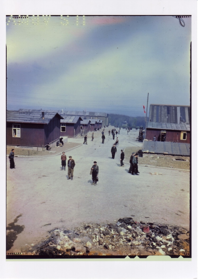 You can see liberated prisoners walking in small groups or individually along the camp street. To the right and left of the camp road, the one-story wooden prisoner barracks can be seen. On the left side, the stone barracks tower above them in the background.