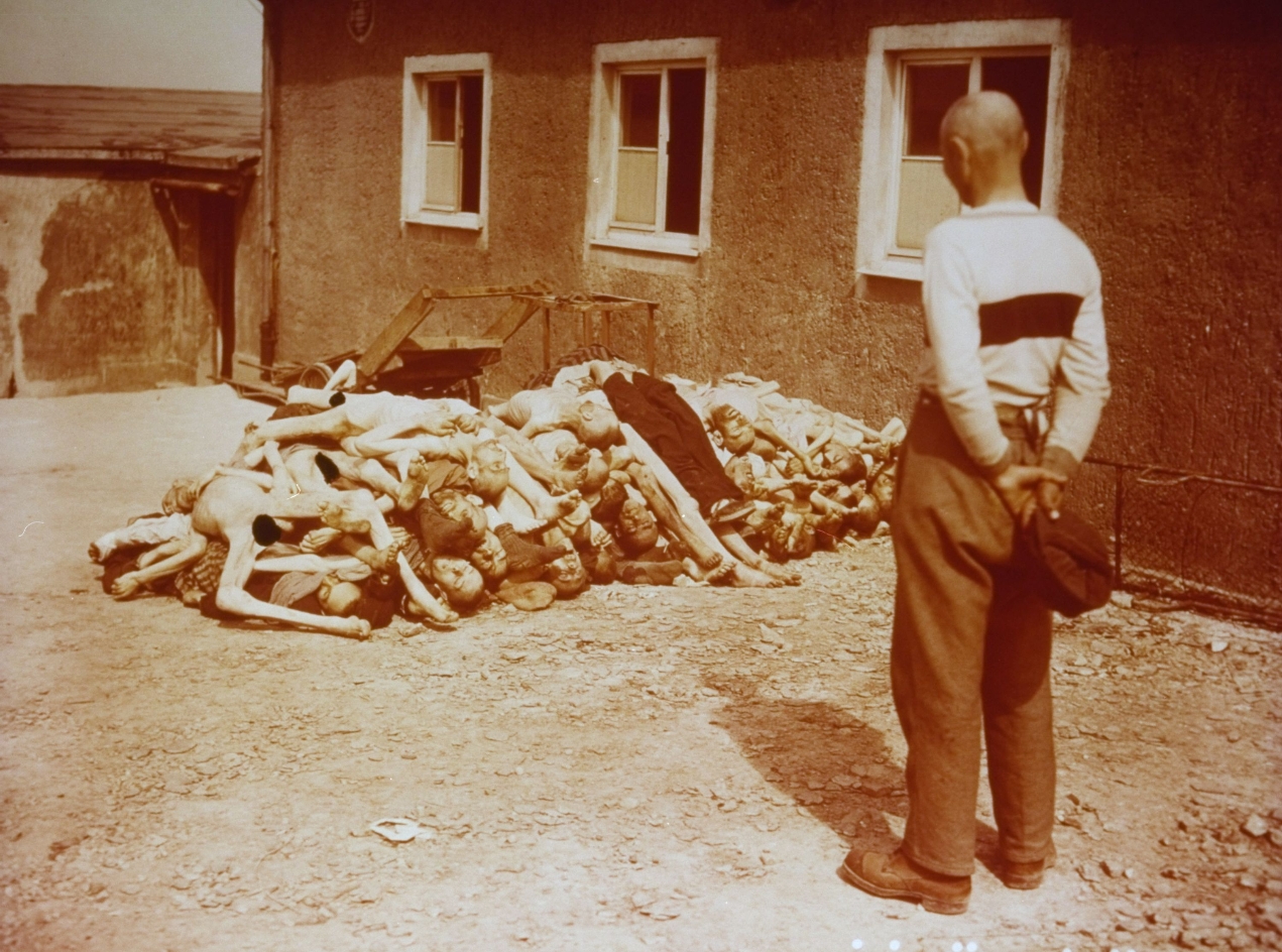 A man in civilian clothes who has taken off his hat in a respectful gesture and holds it behind him. He looks at a pile of emaciated corpses lying in front of him.